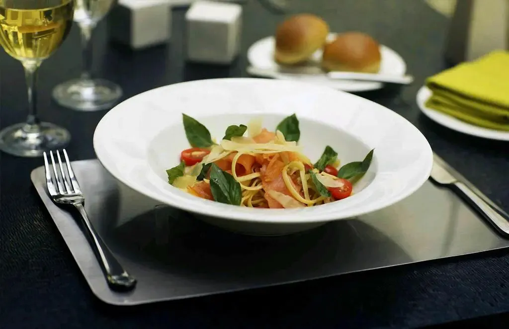 Une assiette de pâtes est présentée sur une table sombre avec des couverts et une boisson.