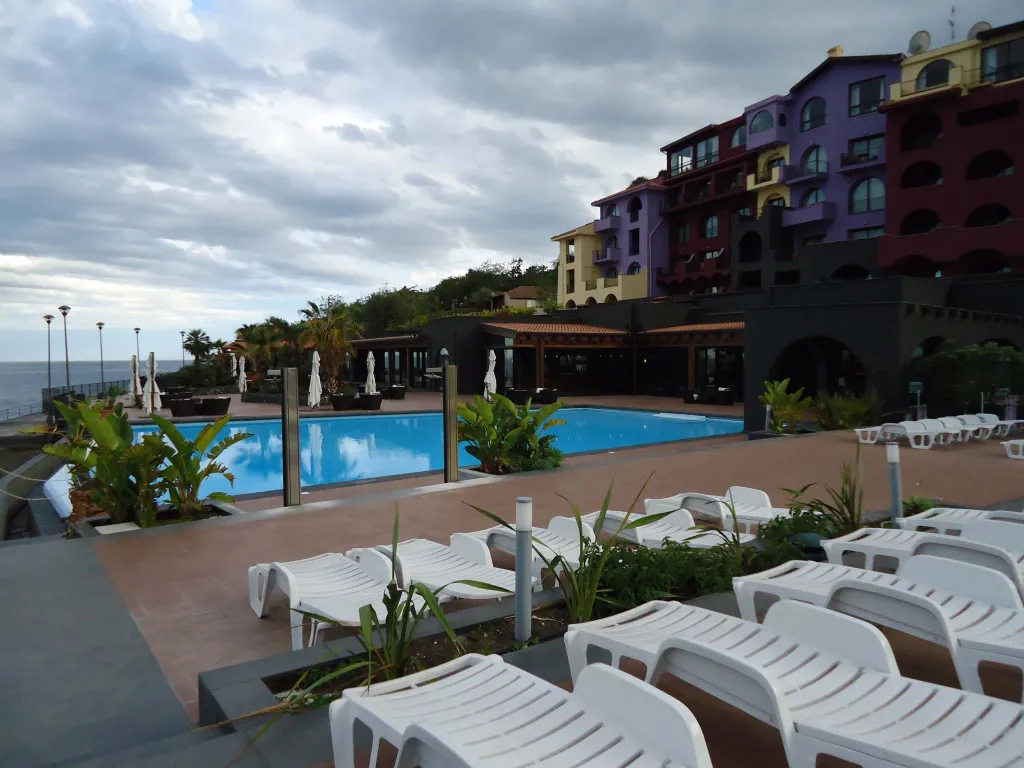 Transats blancs alignés devant une piscine d'hôtel en bord de mer sous un ciel nuageux.