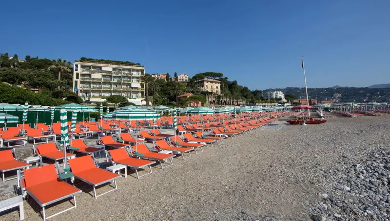 Transats oranges alignés sur une plage de galets face à des bâtiments.