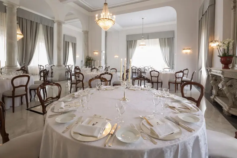 Une salle lumineuse est aménagée avec des tables rondes nappées et des chaises en bois.