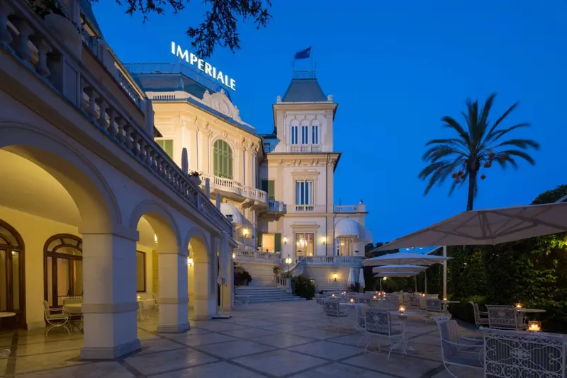 Façade d'un bâtiment illuminé avec une terrasse et des palmiers sous un ciel bleu foncé.