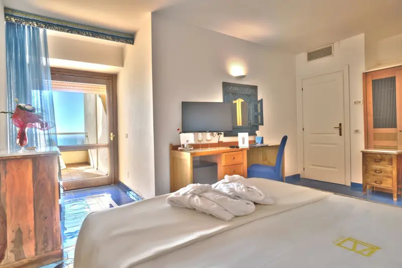 Une chambre lumineuse avec un lit fait et un bureau près d'une fenêtre ouverte sur la mer.