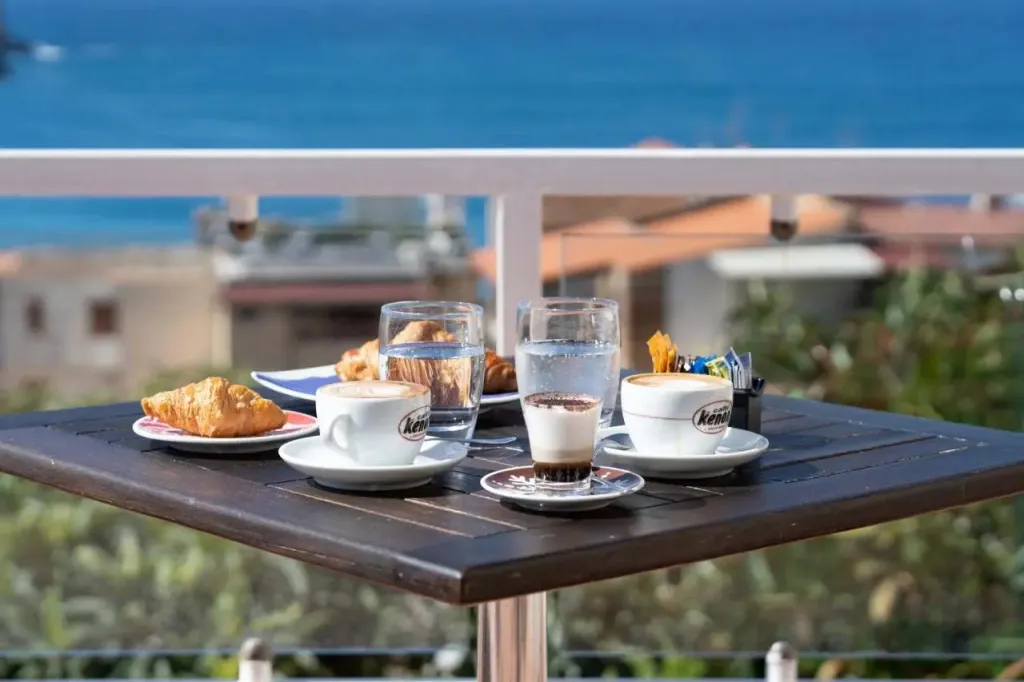 Une table en bois présente un petit-déjeuner avec des viennoiseries et des boissons, face à une vue sur la mer.