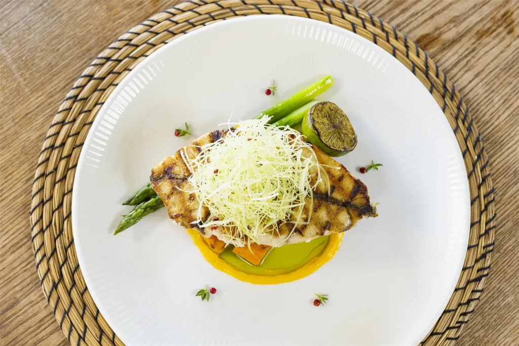 Assiette de poisson grillé agrémentée d'asperges et d'une sauce colorée.