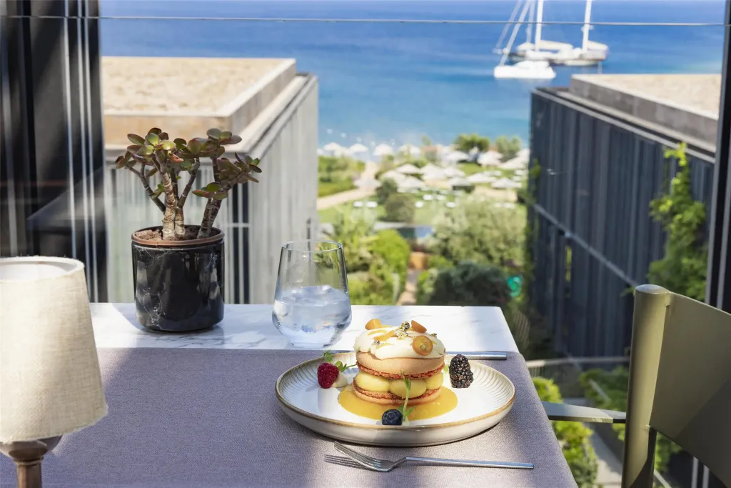 Une table est dressée avec un dessert et une boisson, face à une vue sur la mer et une plage.