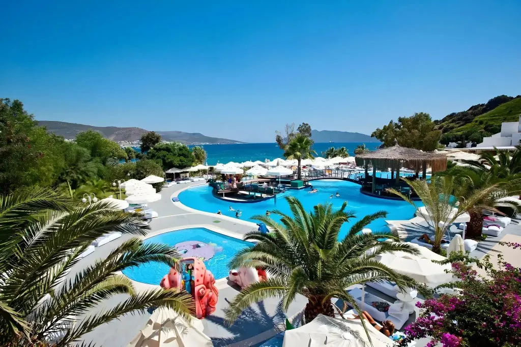 Une piscine turquoise bordée de transats blancs et de palmiers, avec une vue sur la mer.