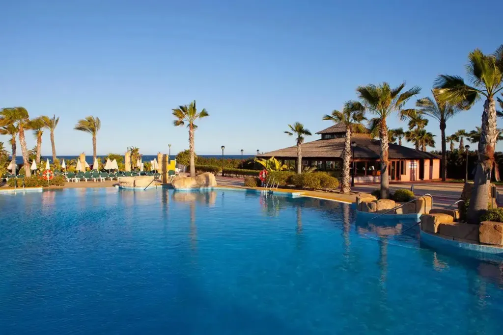 Une piscine bleue bordée de palmiers et de transats sous un ciel clair.