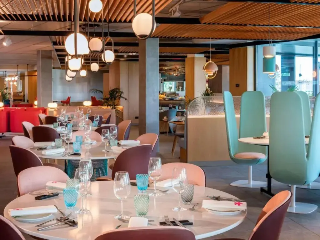 Une salle lumineuse est aménagée avec des tables rondes et des chaises colorées.