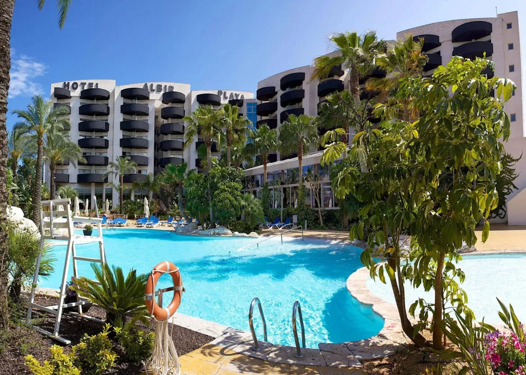 Une piscine turquoise bordée de transats et de palmiers devant un bâtiment hôtelier.