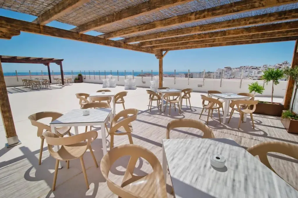 Terrasse ombragée avec tables et chaises donnant sur la mer et une ville côtière.