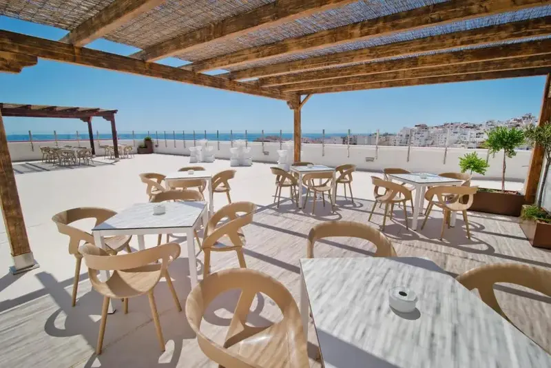 Terrasse ombragée avec tables et chaises donnant sur la mer et une ville côtière.