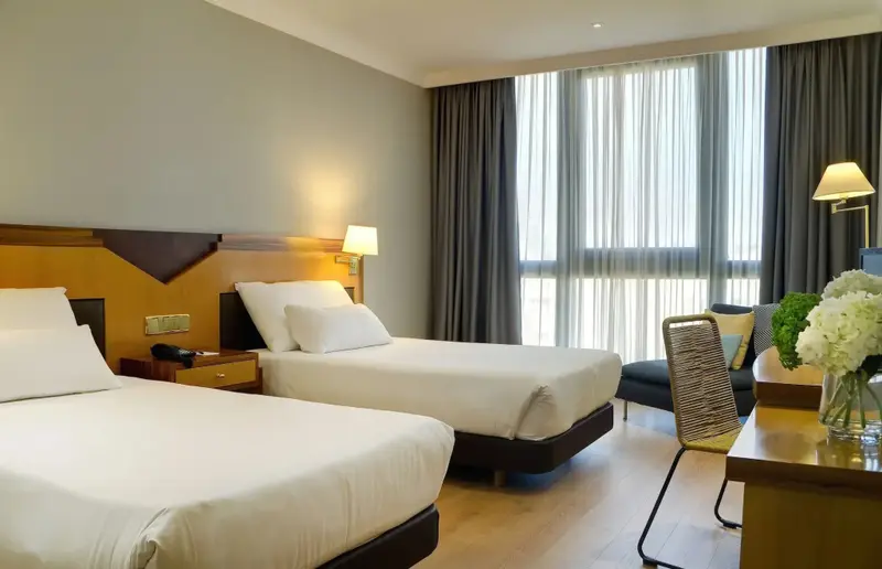 Une chambre lumineuse avec deux lits et un bureau près d'une grande fenêtre.