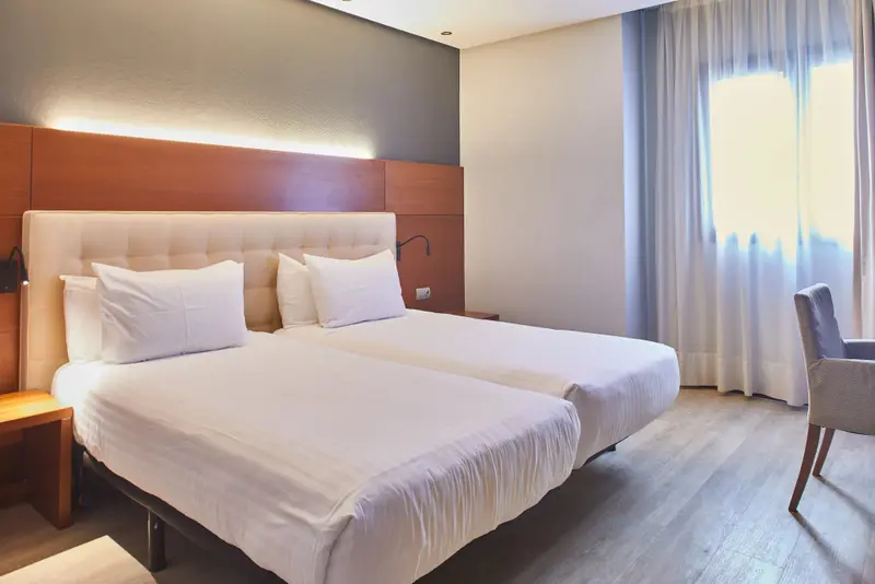 Une chambre lumineuse avec deux lits simples et un fauteuil près d'une fenêtre.