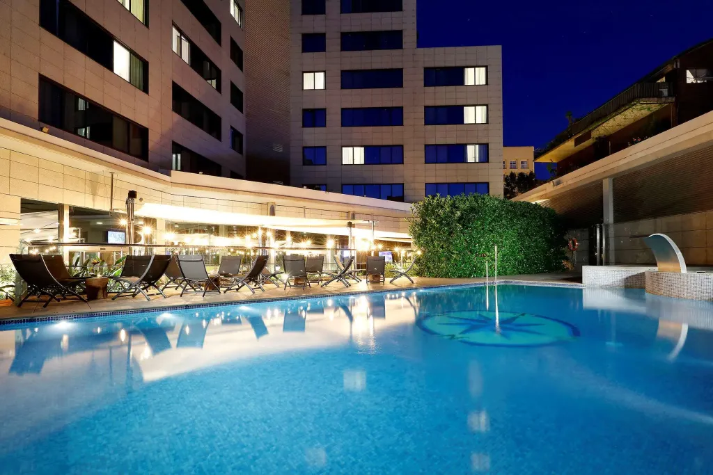 Une piscine illuminée avec des transats et des bâtiments en arrière-plan.