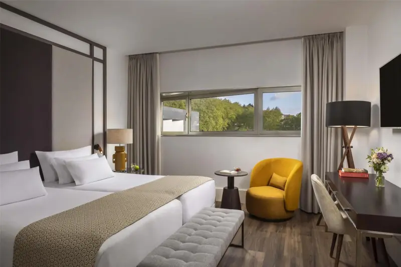 Une chambre lumineuse avec un grand lit et un fauteuil jaune près d'une fenêtre.