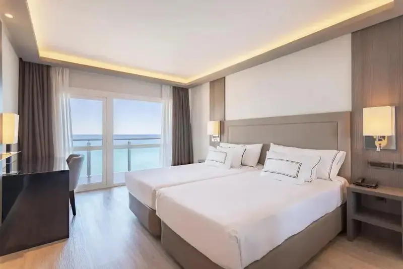 Chambre lumineuse avec deux lits et vue sur la mer depuis un balcon.
