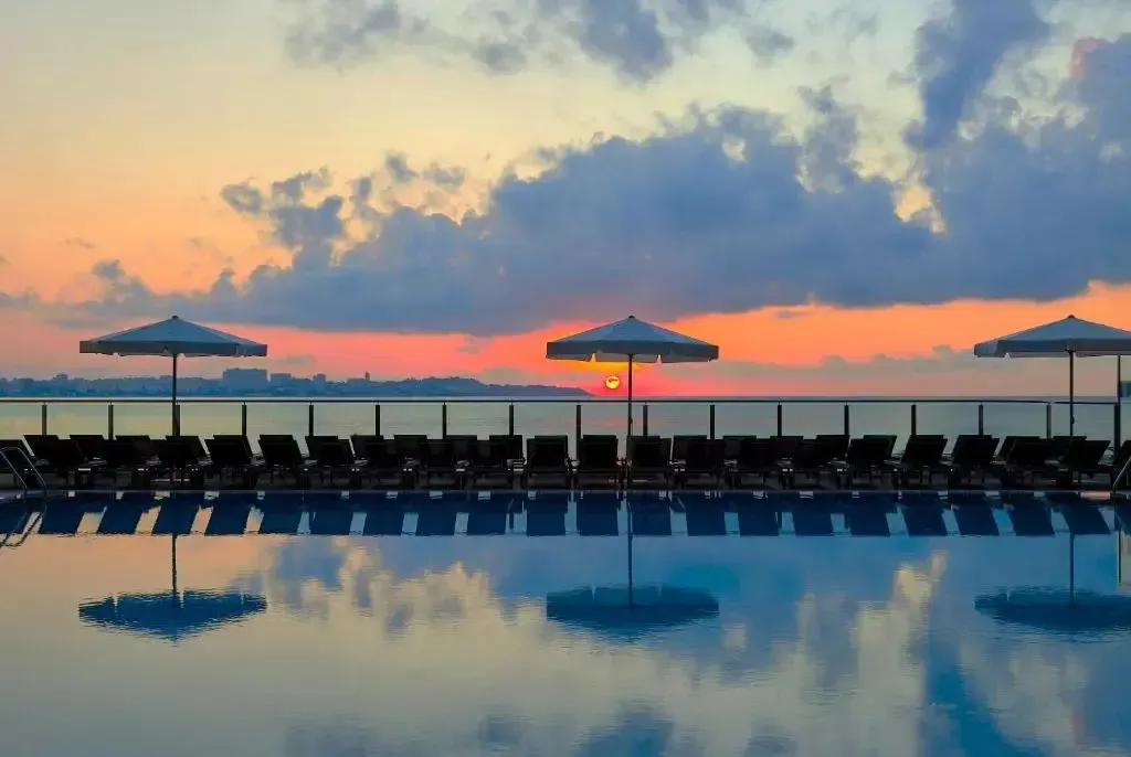 Transats alignés au bord d'une piscine face à la mer au coucher du soleil.