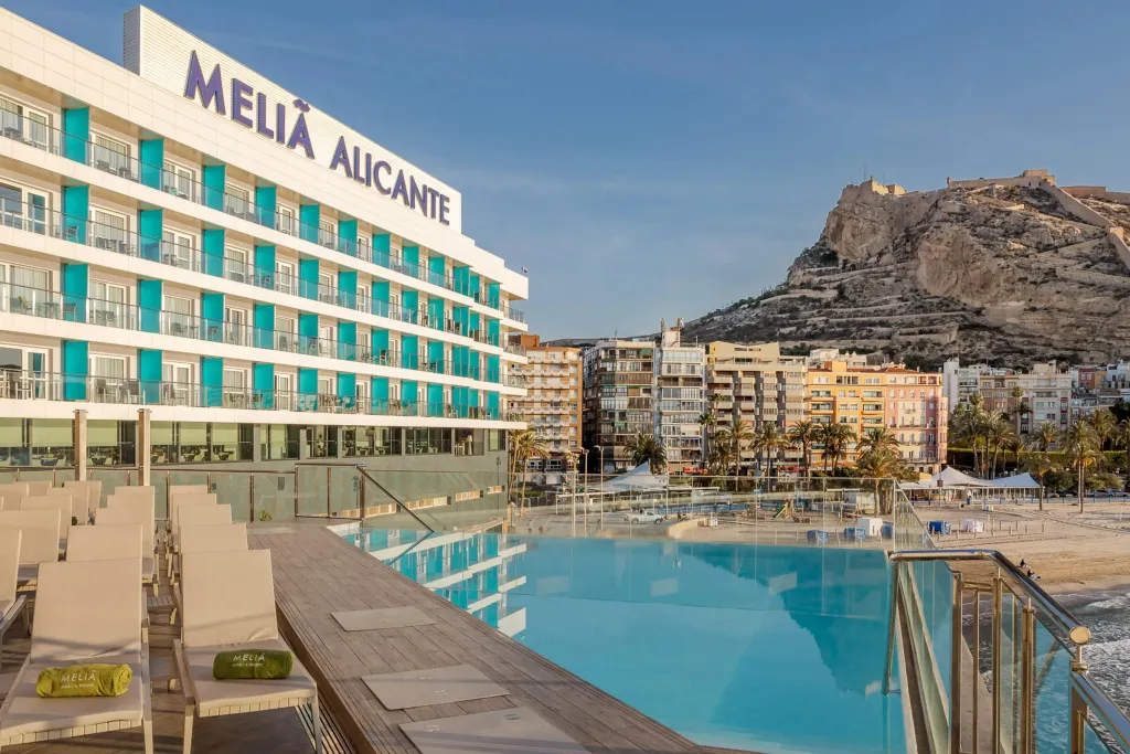 Une piscine d'hôtel avec des transats en bord de mer, devant un bâtiment moderne et une promenade.