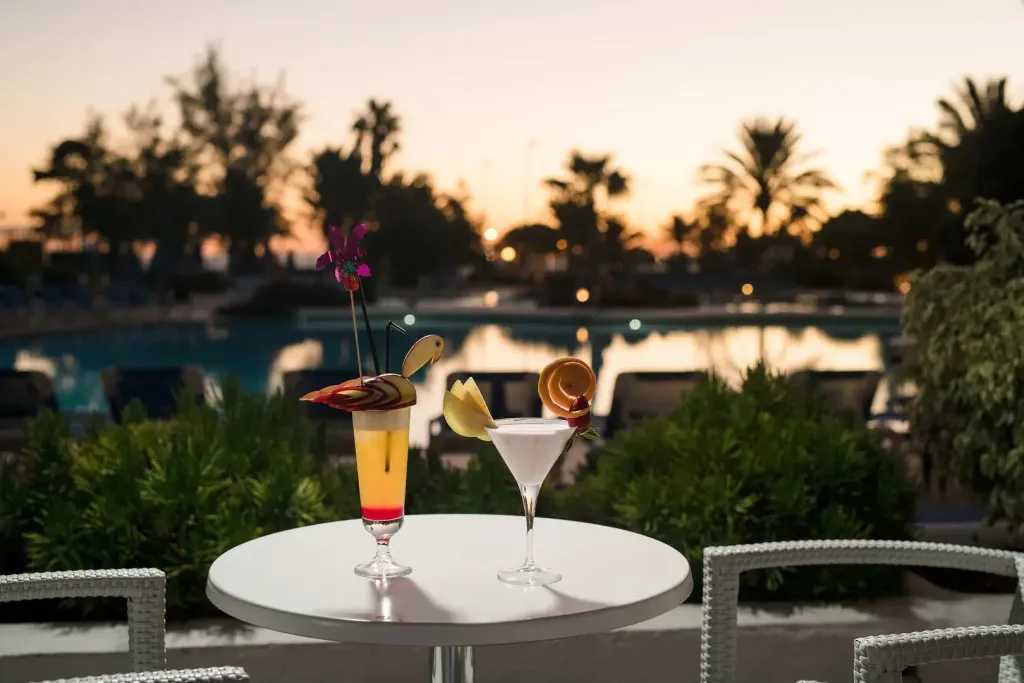 Deux cocktails colorés sont présentés sur une table ronde près d'une piscine au coucher du soleil.