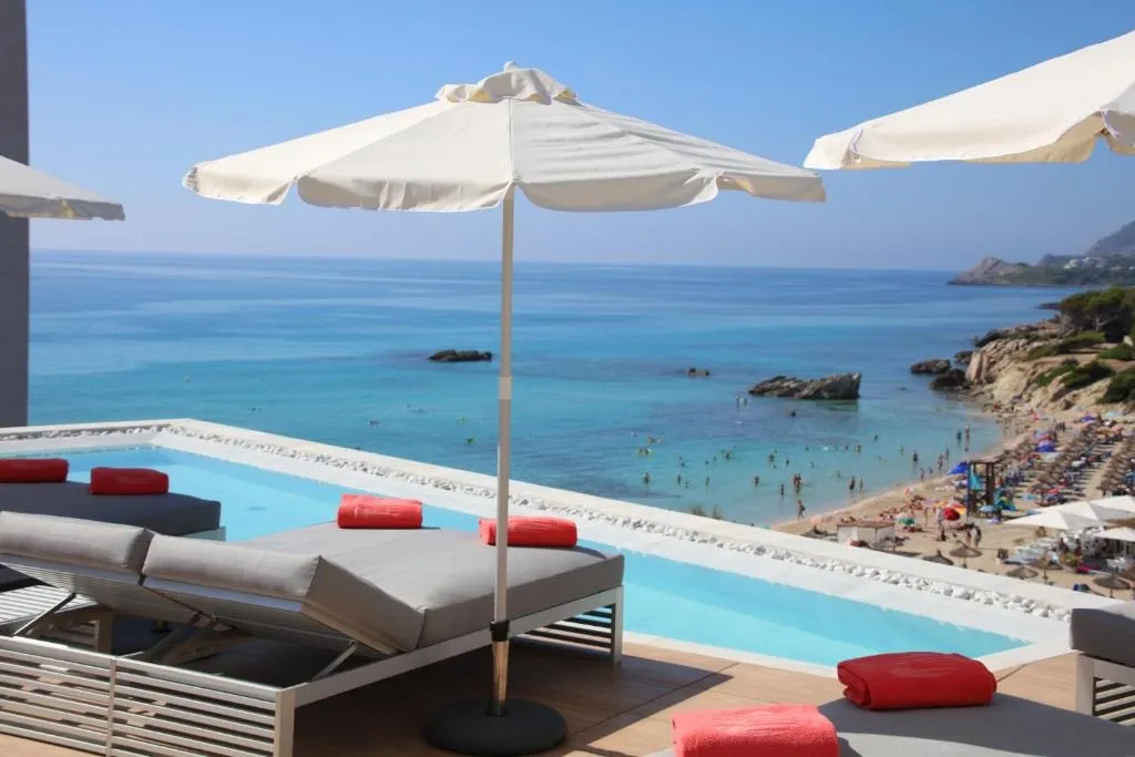 Transats blancs et parasols au bord d'une piscine turquoise surplombant une plage de sable.