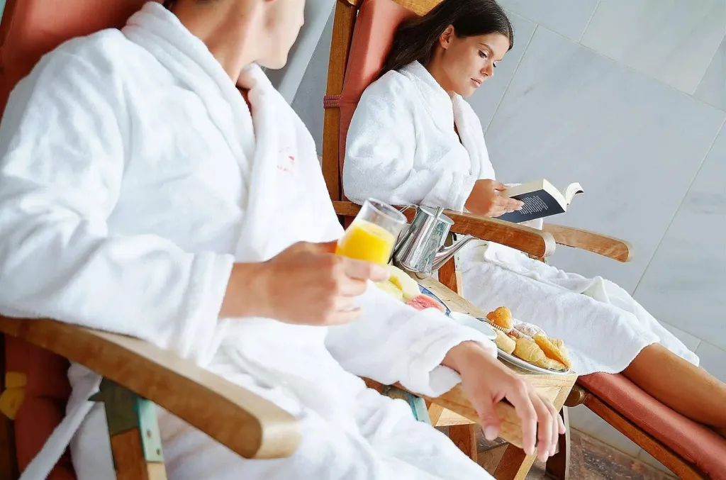 Deux personnes en peignoir se détendent sur des fauteuils en osier avec des viennoiseries et du jus d'orange.