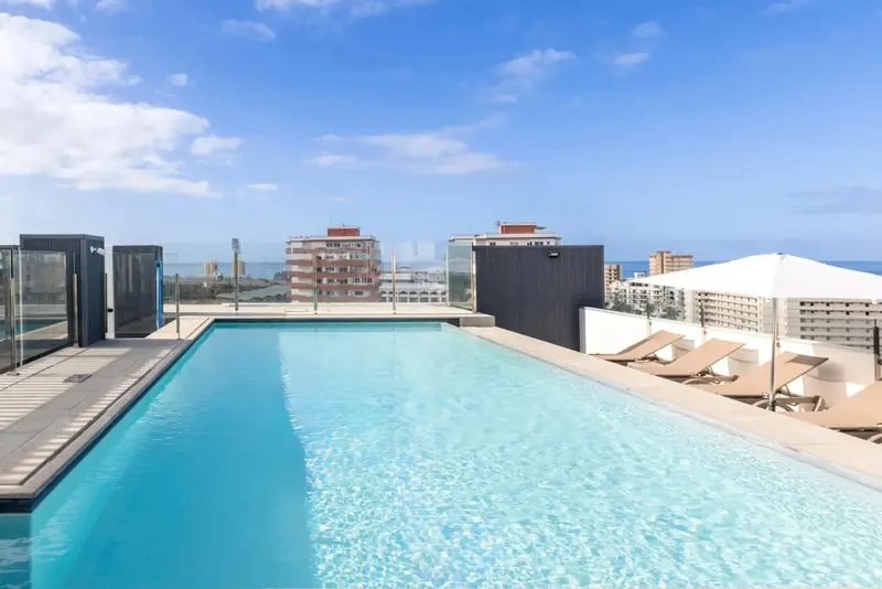 Piscine rectangulaire sur un toit-terrasse avec transats et vue sur la ville et la mer.