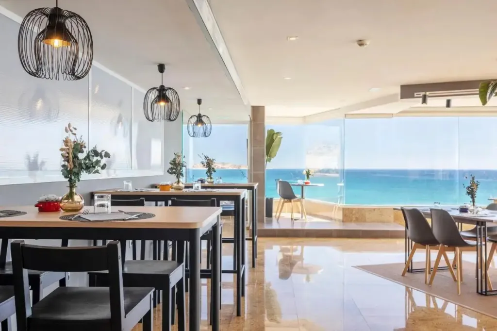 Une salle lumineuse avec des tables et des chaises près d'une grande baie vitrée donnant sur la mer.