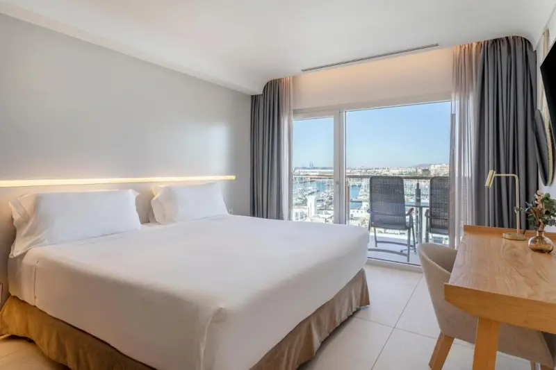Une chambre lumineuse avec un grand lit et un bureau donnant sur un port de plaisance.