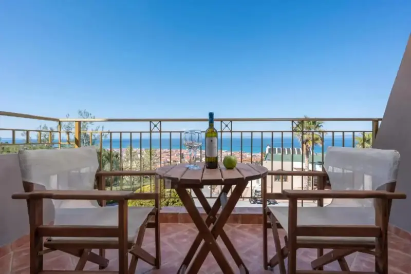 Une table ronde en bois avec une bouteille de vin et des fruits sur un balcon avec vue sur la mer.