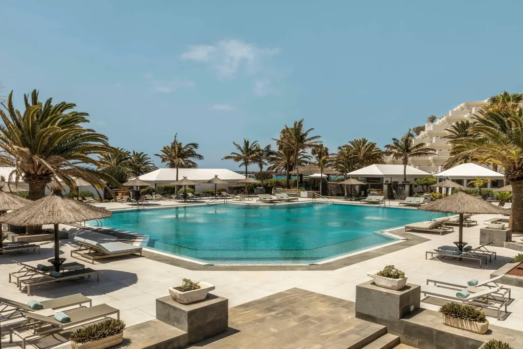 Une piscine turquoise bordée de transats et de palmiers sous un ciel bleu.