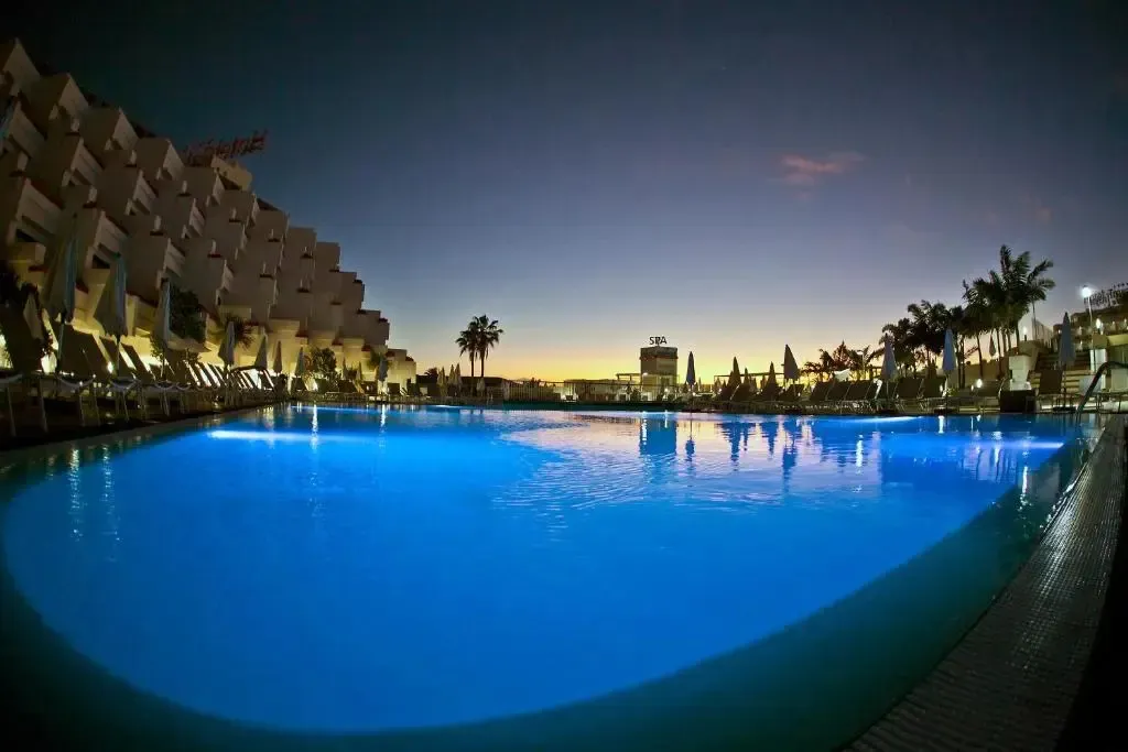 Une piscine illuminée au crépuscule, bordée de transats et surplombée d'un bâtiment à l'architecture particulière.