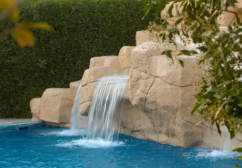 Une cascade artificielle se déverse dans une piscine bleue entourée de végétation.