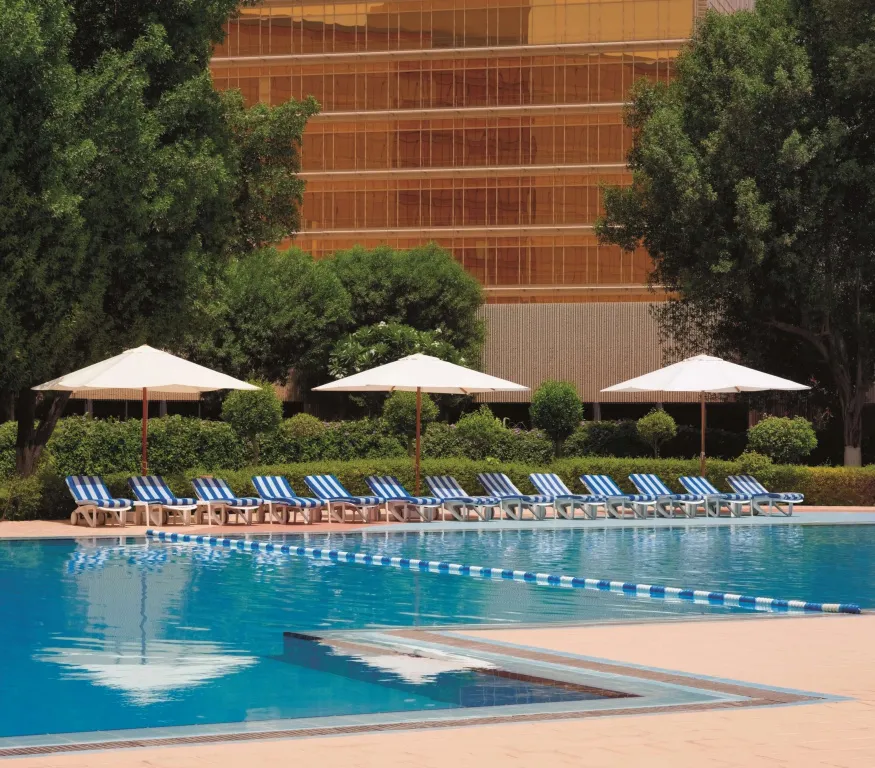 Une piscine rectangulaire bordée de transats blancs et bleus, avec un bâtiment en arrière-plan.