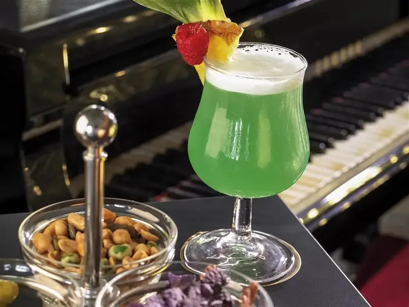 Un cocktail vert est servi avec des cacahuètes et une décoration fruitée devant un piano à queue.