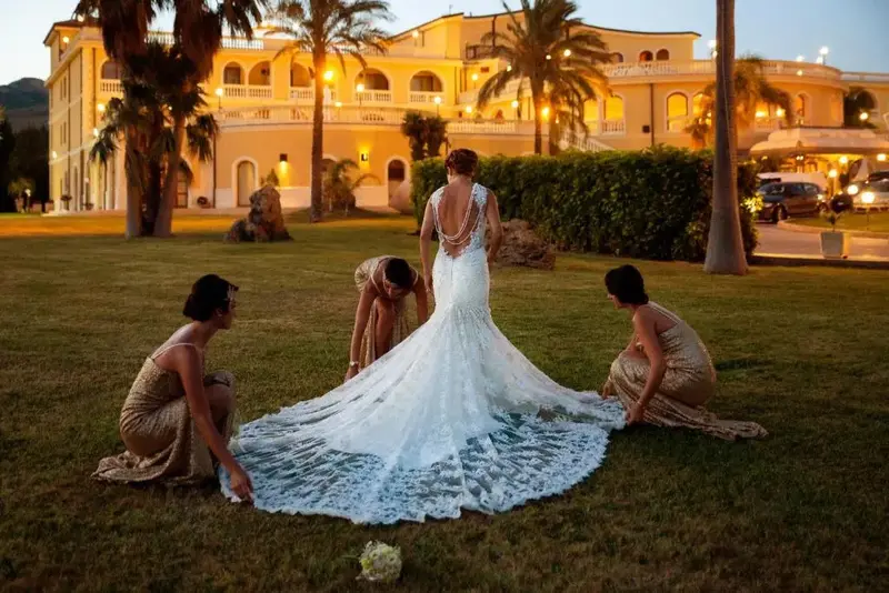 Une mariée et ses demoiselles d'honneur ajustent la longue traîne de sa robe sur une pelouse verdoyante devant un bâtiment illuminé.