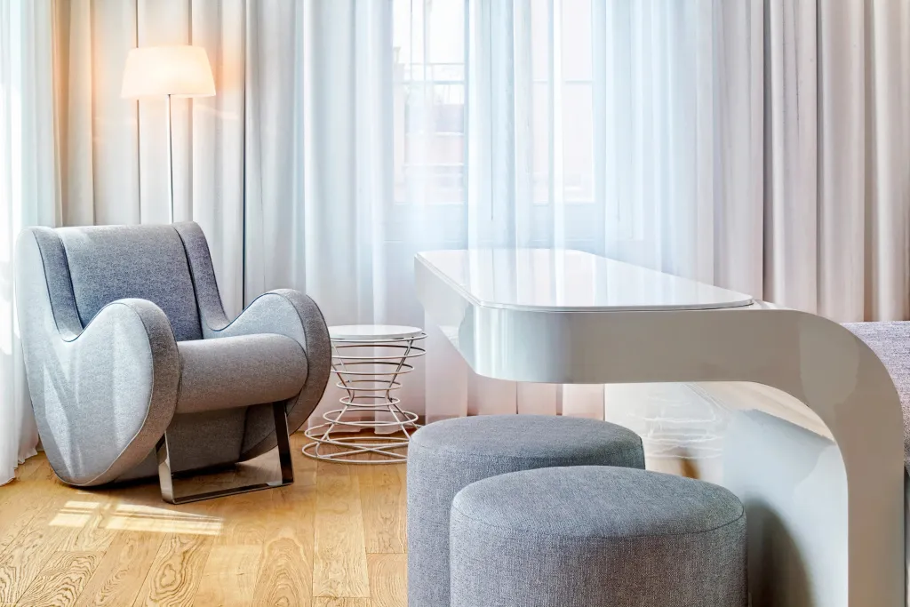 Un fauteuil et des poufs gris reposent devant une fenêtre drapée de rideaux blancs.