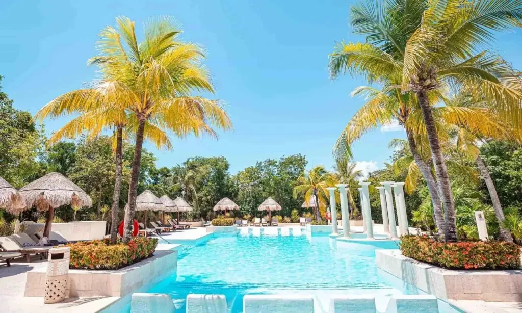 Une piscine turquoise bordée de chaises longues et de palmiers sous un ciel bleu.