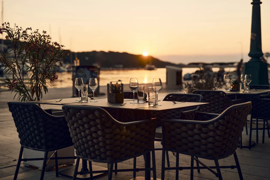 Une table est dressée en extérieur avec des couverts et des verres, face à la mer au coucher du soleil.