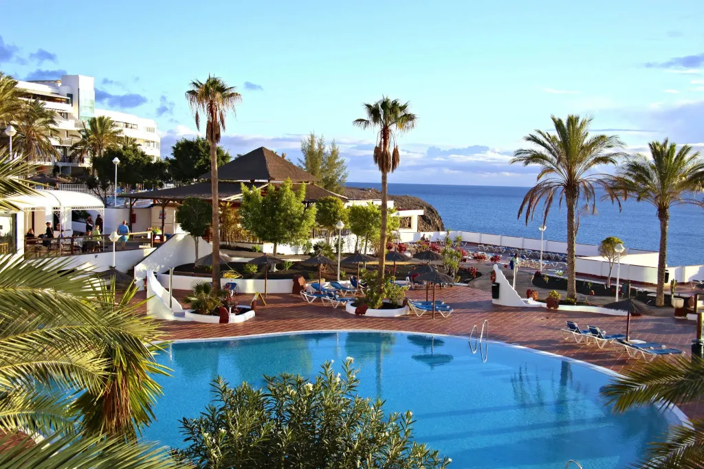 Une piscine ensoleillée avec des transats et des palmiers, surplombant la mer.