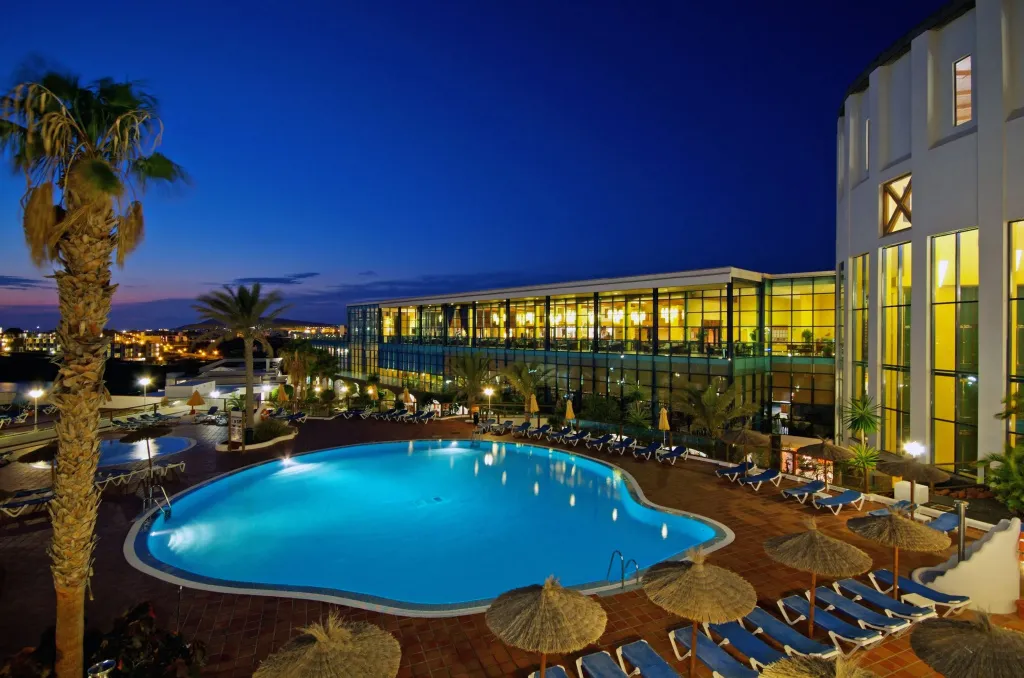 Piscine illuminée d'un hôtel au crépuscule, avec des transats et des palmiers.