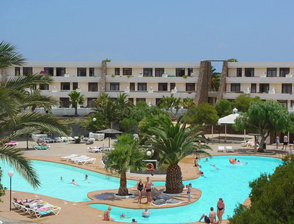 Une piscine ensoleillée avec des transats et des palmiers devant un bâtiment beige.