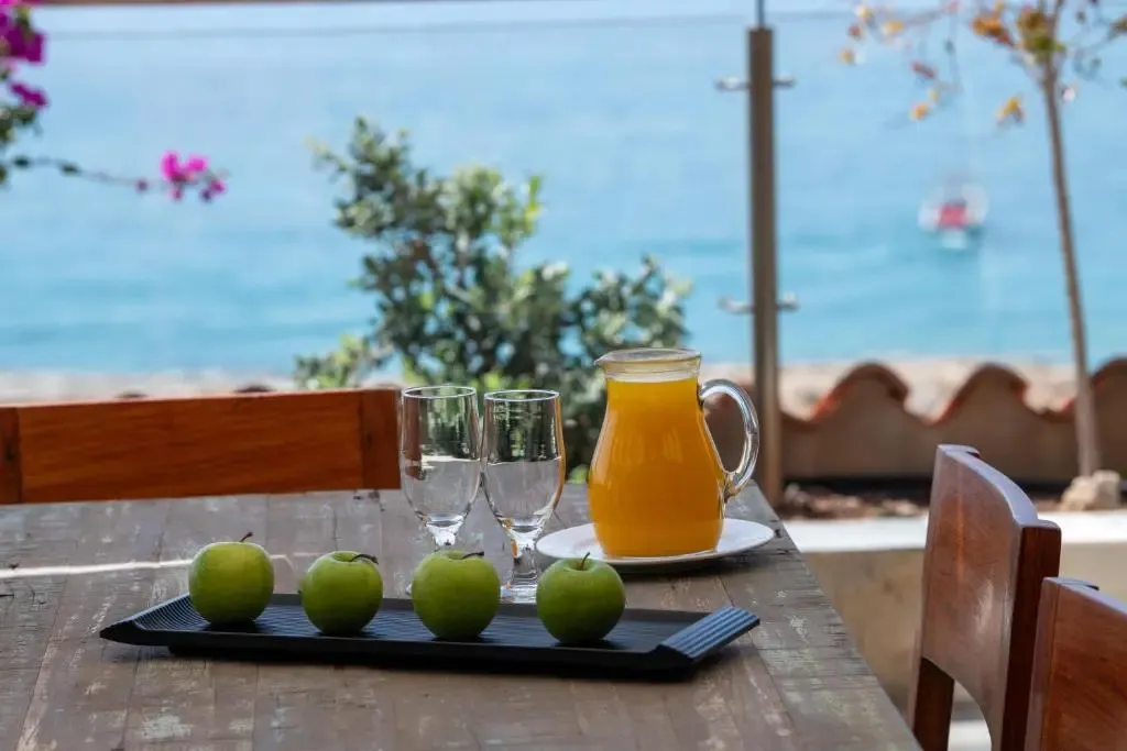 Une table en bois est dressée avec des verres, des pommes vertes et une carafe de jus d'orange, face à la mer.