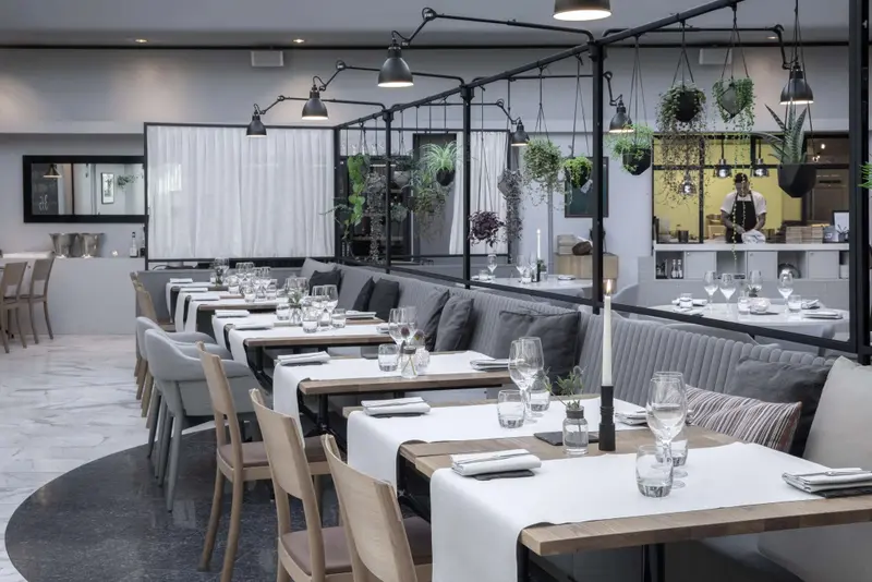 Une salle de restaurant lumineuse avec des tables dressées et des suspensions végétales.