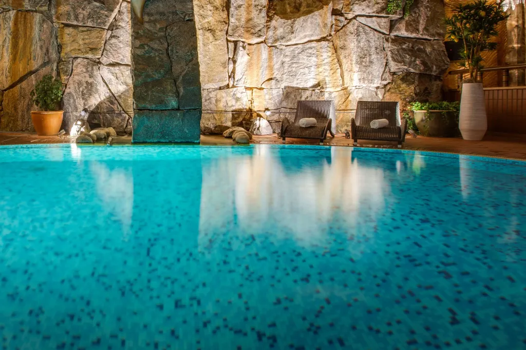 Piscine turquoise avec transats en bois au bord d'un mur en pierre éclairé.