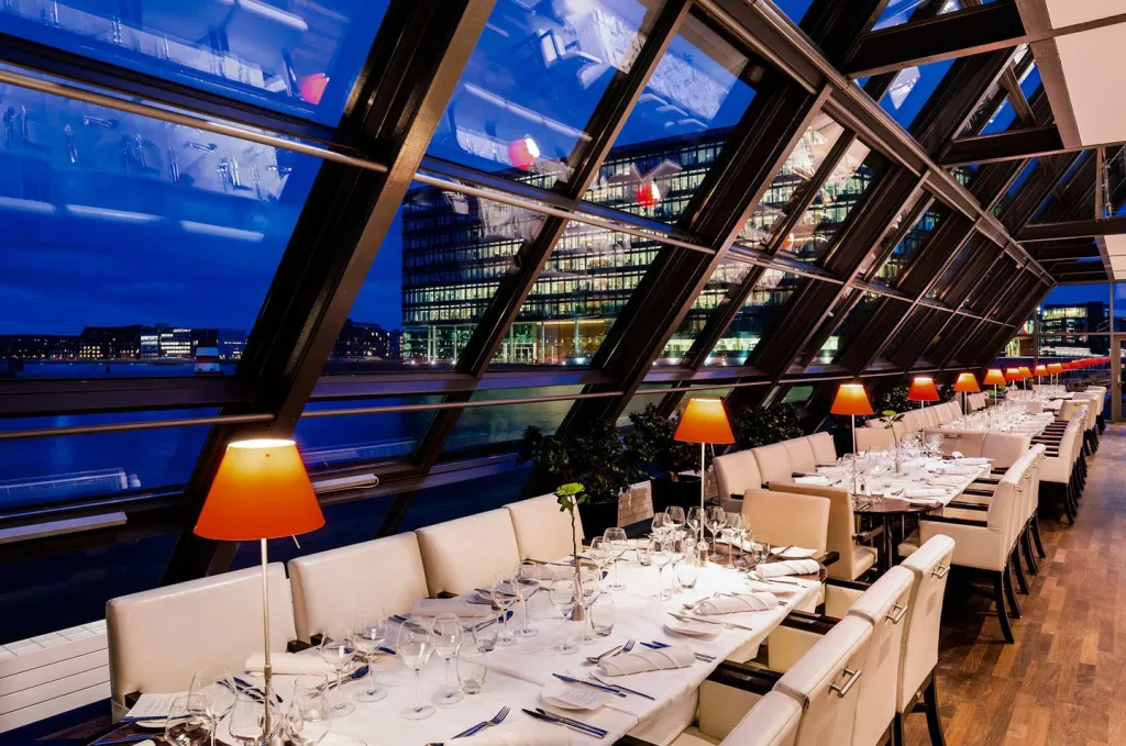 Une salle lumineuse avec des tables dressées face à une vue urbaine nocturne.
