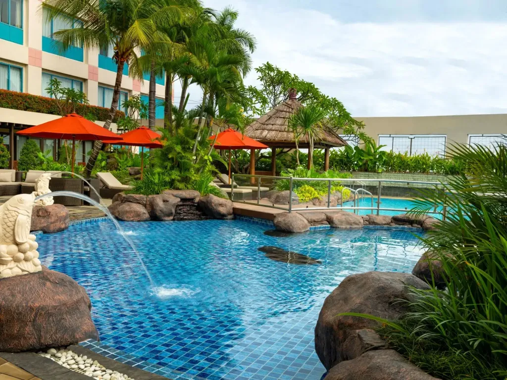Une piscine bleue avec des rochers et une cascade, entourée de végétation tropicale et de parasols.