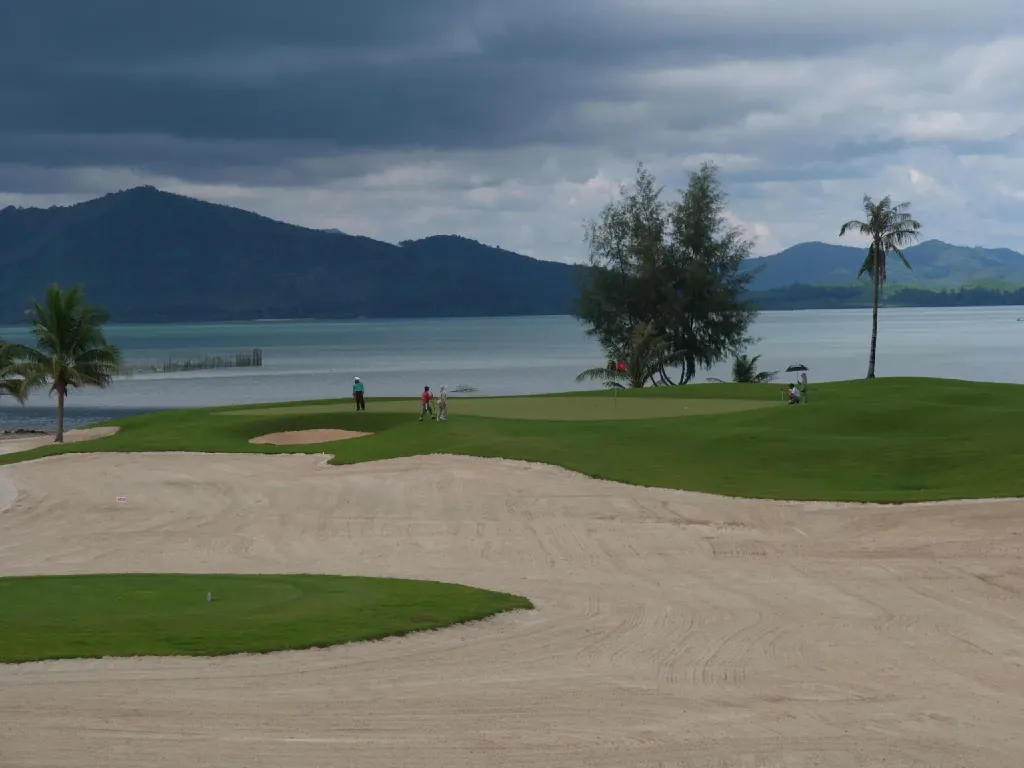 Un terrain de golf verdoyant avec des joueurs et des bunkers de sable, face à la mer et des montagnes.