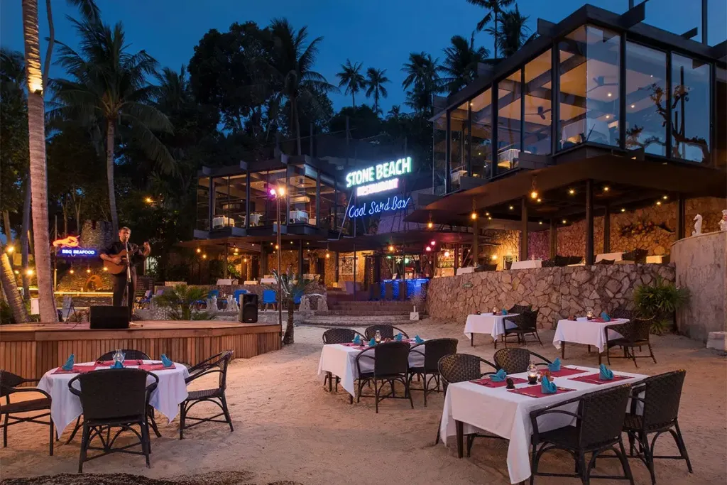 Tables dressées sur le sable devant un bâtiment illuminé au crépuscule.