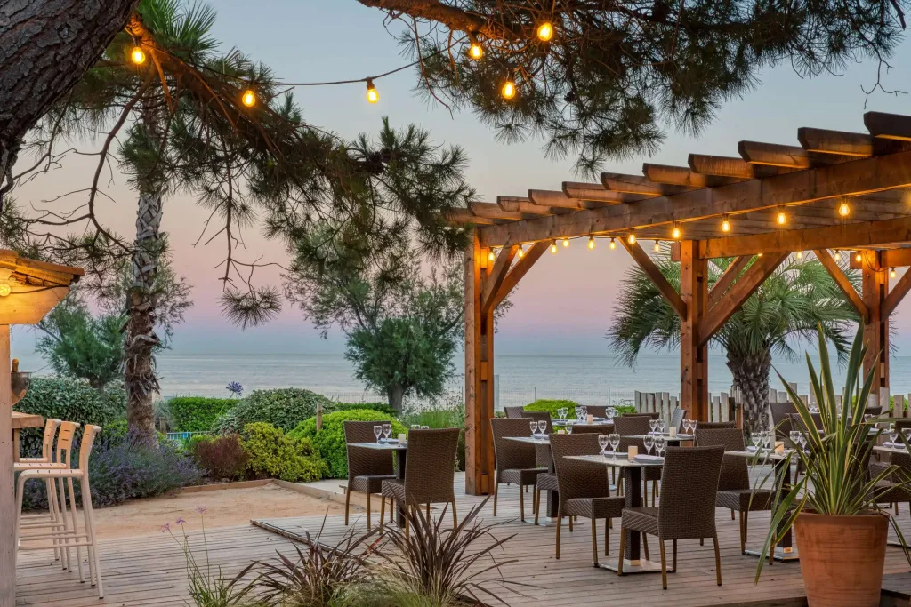 Terrasse ombragée avec tables et chaises face à la mer au coucher du soleil.
