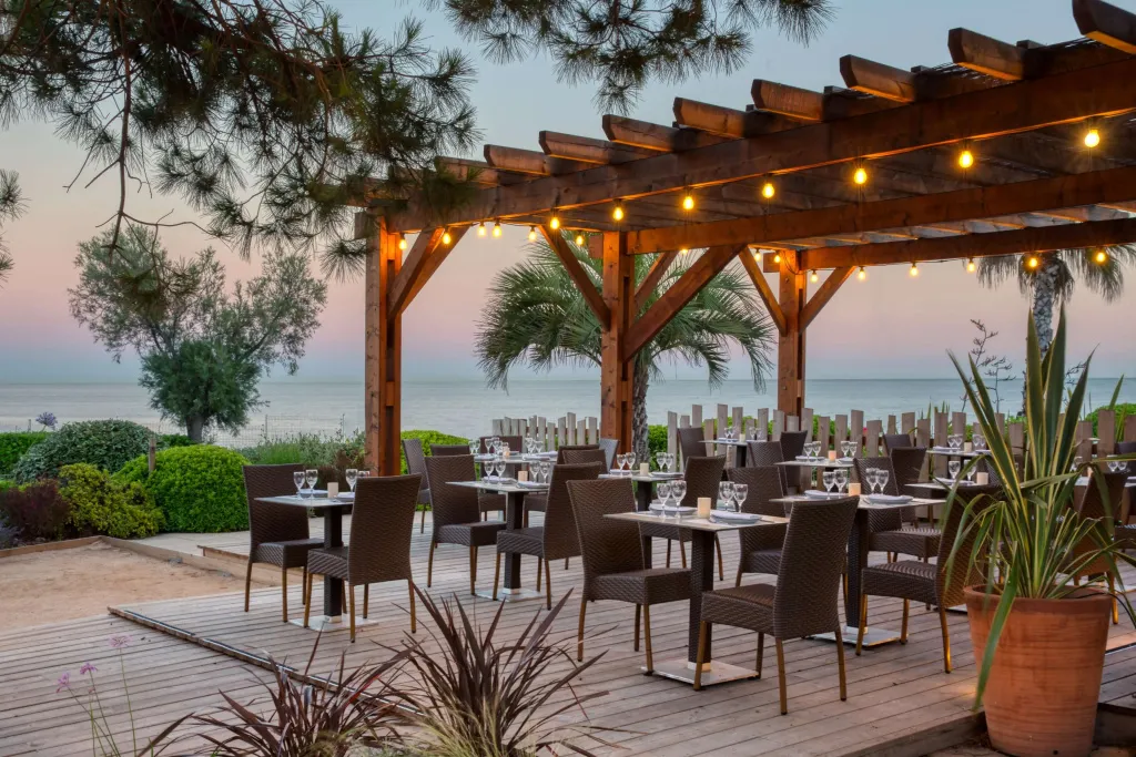 Terrasse ombragée avec tables dressées face à la mer au coucher du soleil.